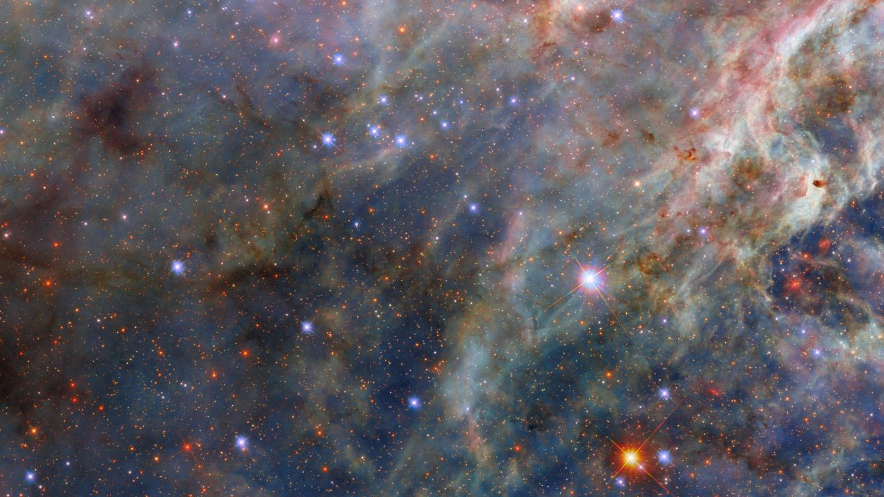 A nebula. The top-left is dense with layers of fluffy pink and greenish clouds. Long strands of green clouds stretch out from here; a faint layer of translucent blue dust combines with them to create a three-dimensional scene. A sparse network of dark dust clouds in the foreground adds reddish-black patches atop the nebula. Blue-white and orange stars, from our galaxy and beyond, are spread amongst the clouds.