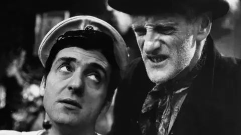 BBC A black and white image of a young man staring up at an older man. The younger man has a white hat and the older man is wearing a neck scarf and black jacket.