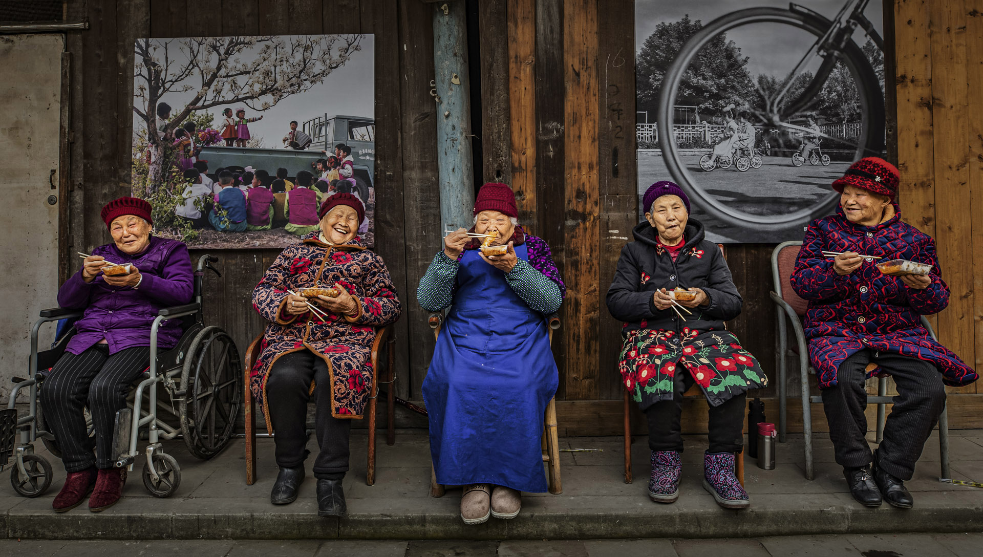 Five elderly women sit in a row on chairs and wheelchairs, enjoying food in colorful attire, against a rustic wooden backdrop
