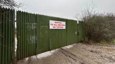 A green metal gate stretches across the entrance to the land. A white sign with red writing reads: "Notice to all driver no tipping." 