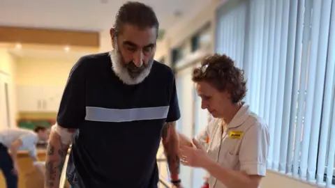 NHFT Anthony Dexter with Marion Packwood. Anthony, has short dark hair and a grey heard, is wearing a black T-shirt, and Marion, is on  hand to his right. They are in a rehab centre. 