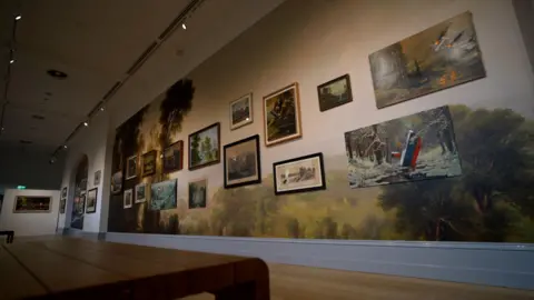 A wide shot showing a wall of artworks in an art gallery. The picture is taken from a wooden bench which can be seen in the foreground.