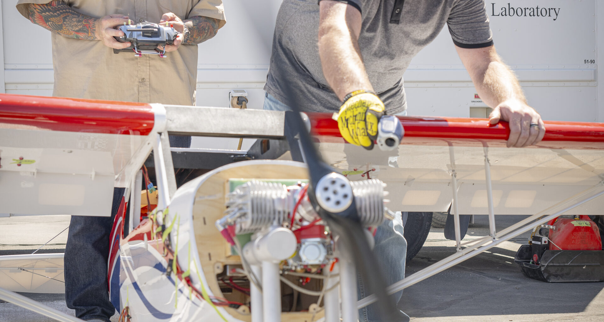 One white man with a long beard and tattoos on his arms holds a silver and gray controller, while another white man leans over to hold a small aircraft in place. The two men are both wearing hearing protection, which look like headphones. The aircraft looks to be only a few feet tall.