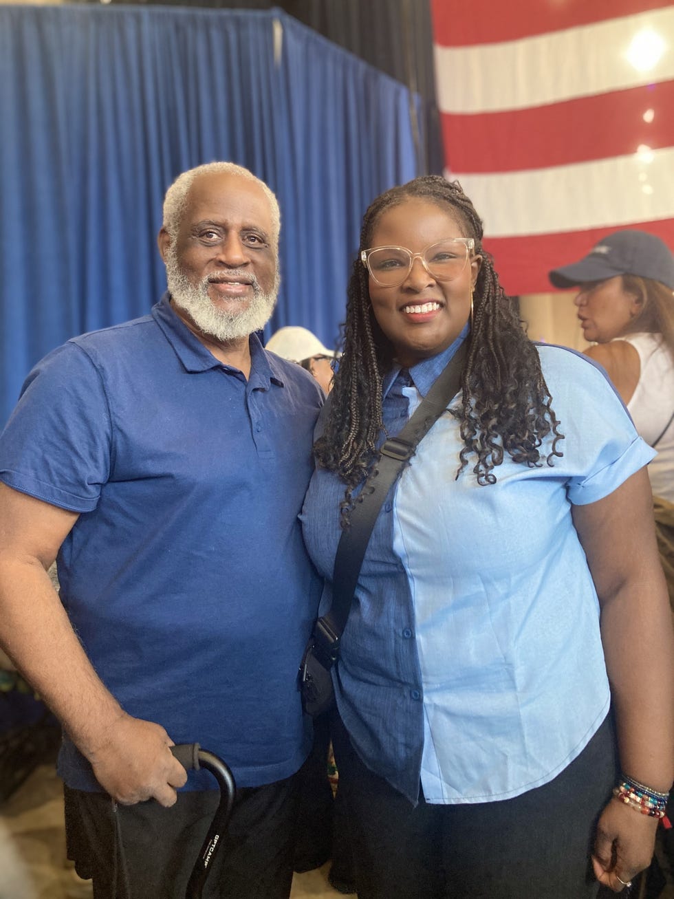 two individuals posing together at an event with a backdrop of a blue curtain and an american flag