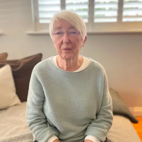 Pauline Collier sitting on a bed. She has short, white hair and is wearing a pale blue jumper.