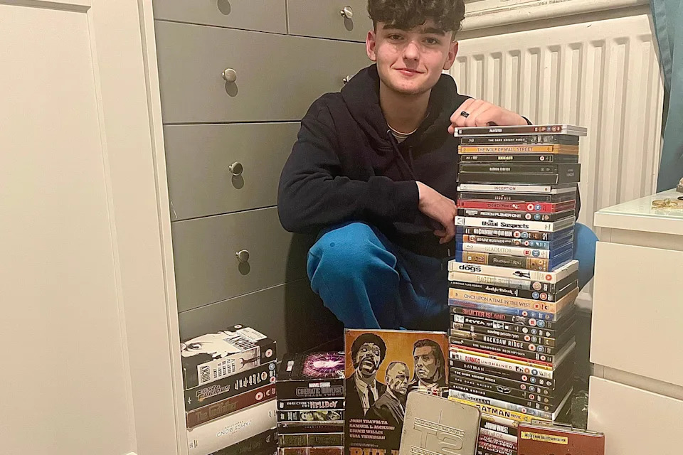 Declan sitting on the floor next to two tall stacks of DVDs and box sets, with additional DVDs arranged in front. The scene includes a grey chest of drawers on the left and a white radiator in the background.