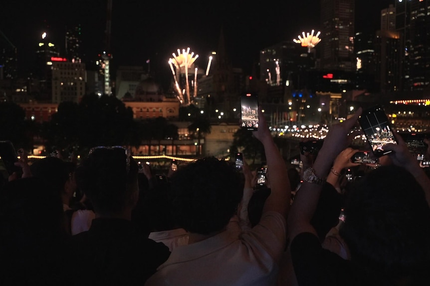 A photo taken behind a crowd, many of whom hold up phones filming fireworks coming from near city buildings at night.