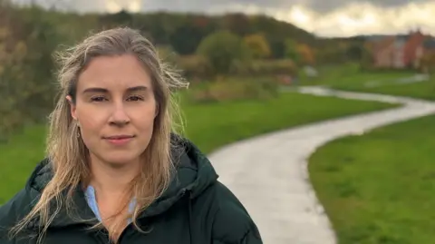 Katie Anderson looks straight at the camera - she has brown hair with blonde highlights and is wearing a dark winter coat. Behind her is a housing estate and a winding footpath flanked by grass on either side.