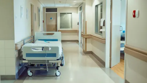Getty Images A portable white hospital bed sits tight against a wall in a bright, empty corridor. Small spotlights shine a pale light on the clean, neutral colour palette of the ward.   