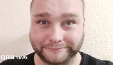 A close-up selfie of a man with a light brown beard and light-brown eyes. He is smiling at the camera. There is a white wall behind him.