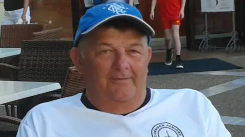 Family handout A man wearing a Cumnock Juniors white sports T-shirt with black sleeves and a blue cap, seated outdoors at a café. Behind him is a building entrance with glass doors, decorative panels, and several people walking nearby.