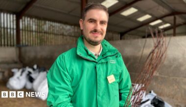 A man with brown hair and facial hair smiles as he stands in a barn, holding a sapling. Large white bags containing saplings can be seen behind him. He is wearing a green, Forestry England branded coat, and a name badge.