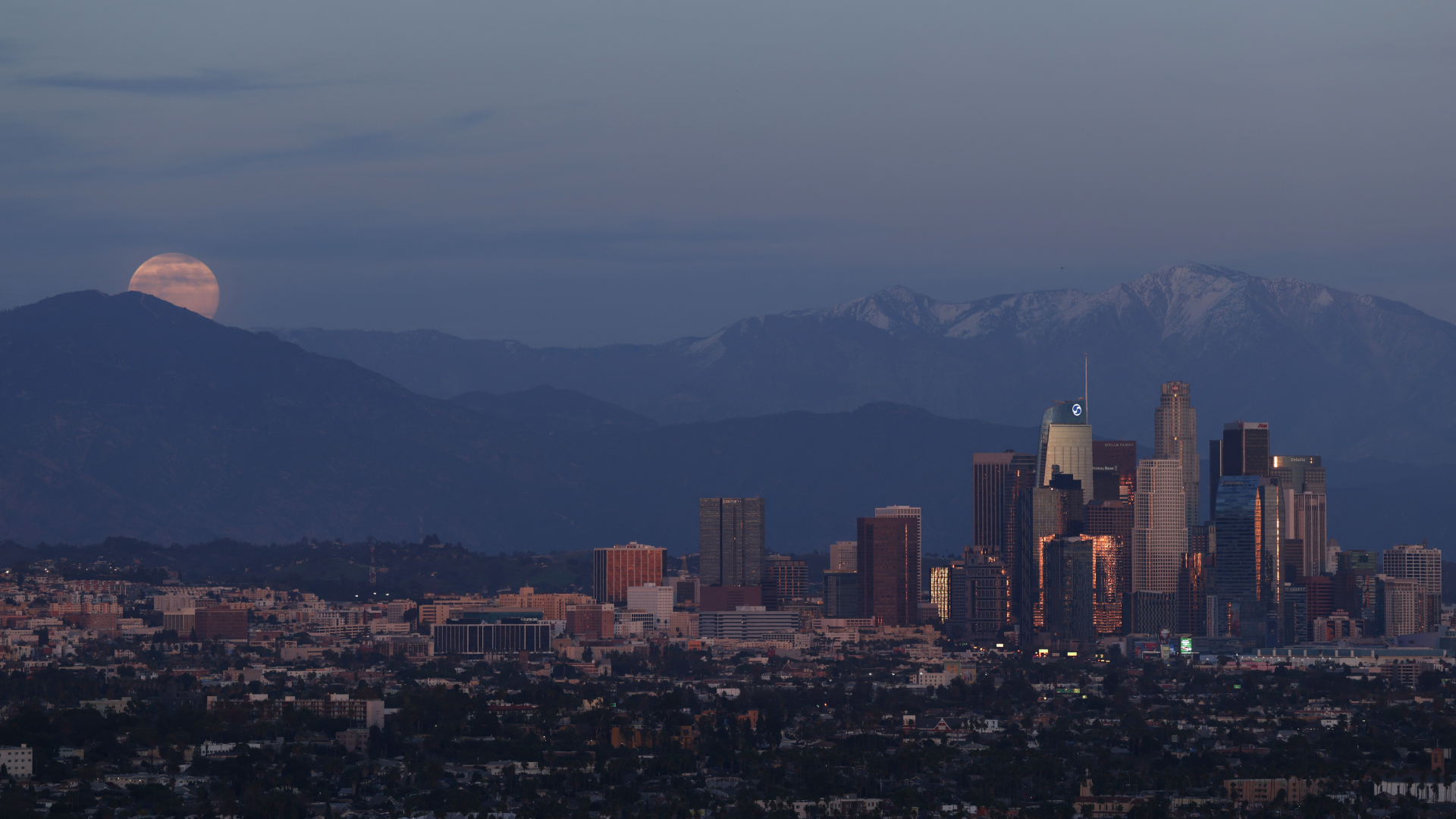 A full moon is pictured rising over a mountain range above a city, as skyscrapers huddle to the left of the shot.