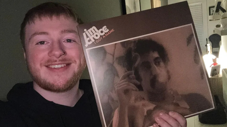 Saul, wearing a black hoodie, is standing holding two vinyl records. The background has green wall with framed artwork and a round mirror.