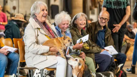Max Willcock A group of people - three women and a man - sit and laugh at a pet blessing service inside a cathedral. There is a small, tan-coloured, scruffy dog sat on the lap of one woman, which is being sniffed by a Basset hound.