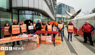 A group of people standing outside a glass building holding orange cards. They say things like "patients need doctors, doctors need jobs" and "doctors need jobs now". They are wearing orange bucket hats and beanies, and have an orange table with more signs leaning against it.