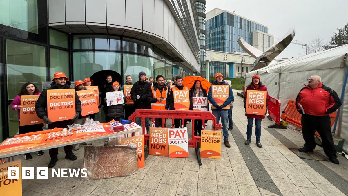 A group of people standing outside a glass building holding orange cards. They say things like "patients need doctors, doctors need jobs" and "doctors need jobs now". They are wearing orange bucket hats and beanies, and have an orange table with more signs leaning against it.