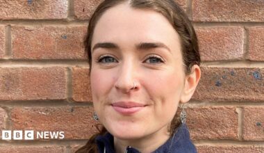 A headshot of a young woman with brown hair tied back. She is standing in front of a brick wall.