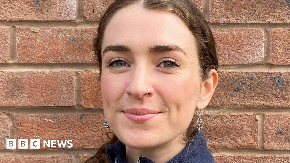 A headshot of a young woman with brown hair tied back. She is standing in front of a brick wall.