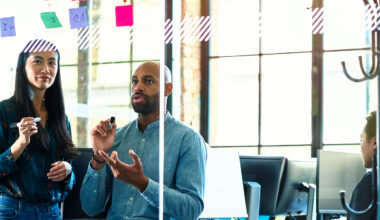 Three colleagues collaborate in a bright office, discussing ideas with sticky notes on a glass wall.