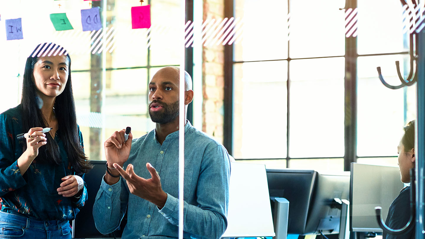 Three colleagues collaborate in a bright office, discussing ideas with sticky notes on a glass wall.