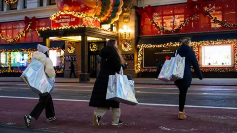 Bloomberg via Getty Images Three shoppers carry large shopping bags while crossing the street, in front of a Macy's store decorated with holiday lights.
