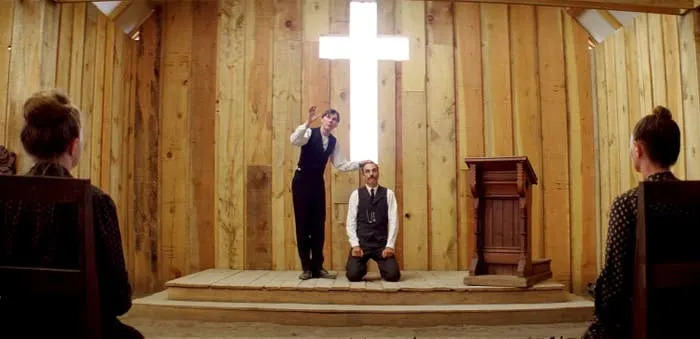 Two individuals inside a rustic wooden church; one standing and gesturing passionately, the other kneeling in front of a bright cross