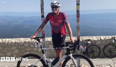 A man wearing cycling gear stands and smiles while standing next to his white bicycle, which he is holding up. Behind him is a brick wall and two posts.