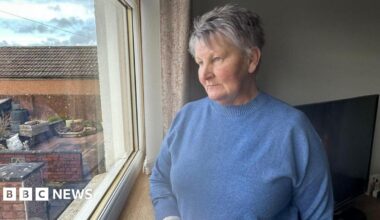 Nancy Dunnachie, who has short, grey hair, is wearing a blue knitted jumper and is staring out of a window