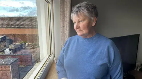 BBC Nancy Dunnachie, who has short, grey hair, is wearing a blue knitted jumper and is staring out of a window