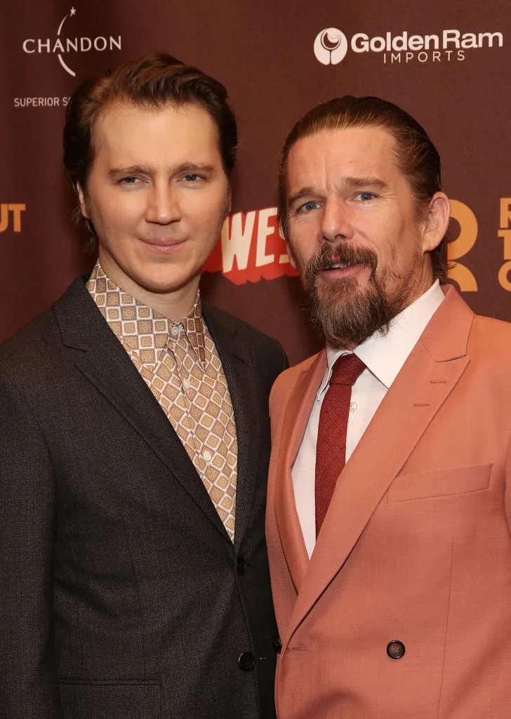 Two men on a red carpet; one in a patterned shirt and dark suit, the other in a peach suit and white shirt with a red tie