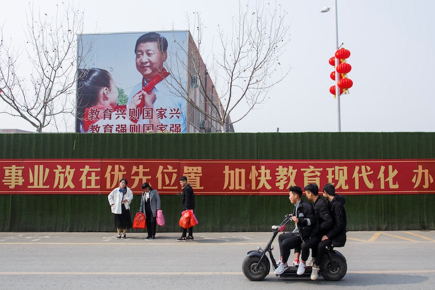 Men ride a scooter past a poster showing Chinese President Xi Jinping on the side of a school 