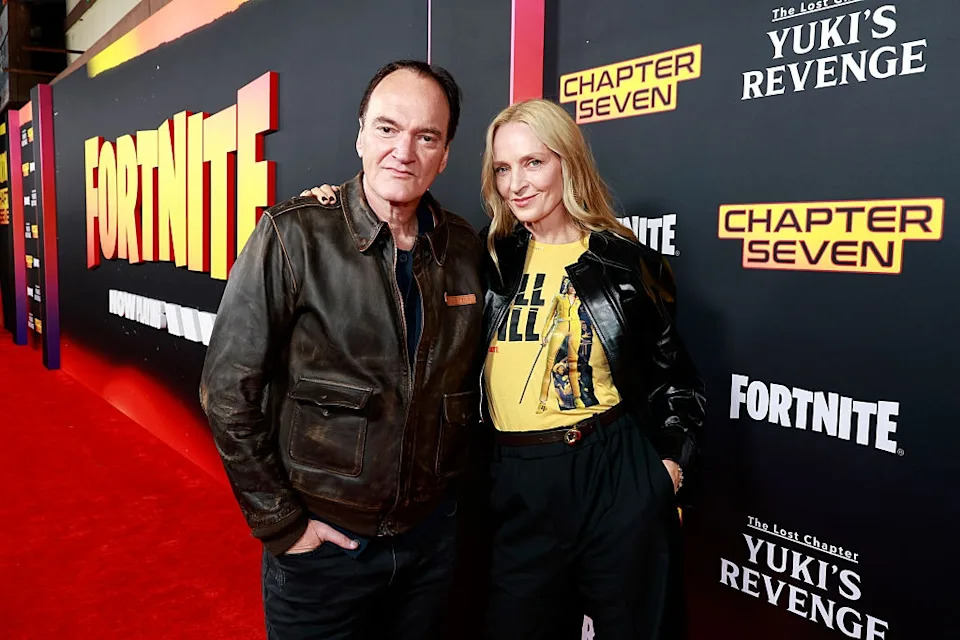 Two people on a red carpet, one in a leather jacket and the other in a graphic tee and leather jacket, posing by "Fortnite" and other logos