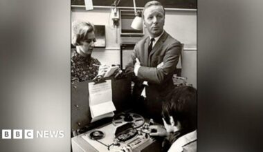 A sharply dressed man in a suit stands with his arms folded whilst a woman takes notes onto a a pad of paper. Another women sits in-front of an old fashioned tape reel player