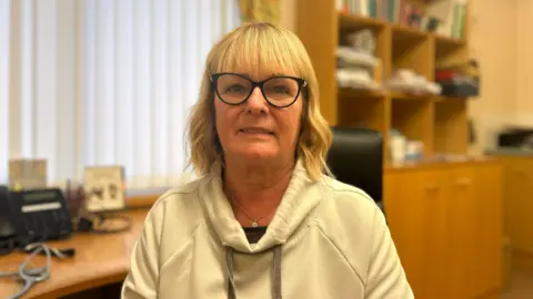 Dr Frances O'Hagan is a woman with short blonde hair, black glasses and is wearing a white hooded top and a necklace. She is sitting in the office of her general practice which is out of focus in the background. A wooden desk, wooden cabinets and window with white blinds.