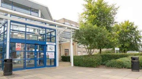 Warrington and Halton Teaching Hospitals NHS Foundation Trust The front entrance of Warrington and Halton Teaching Hospitals NHS Foundation Trust. It is a grey building with a blue front. There is also a tree right outside the entrance.