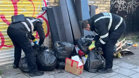 Two environmental officers wearing dark protective clothing look through a number of black plastic refuse bags which are piled against a wall along with other pieces of furniture