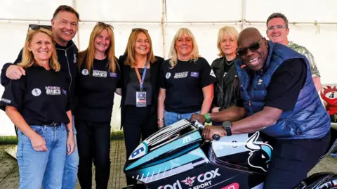 IOM TT Carl Cox sitting on a replica of Michael Dunlop's record-breaking motorbike, which features his logo. Volunteers with the Mannin Cancers Support Group are standing alongside him.