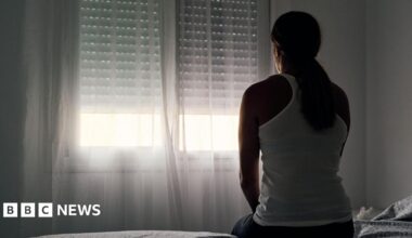A woman wearing a white vest top with her hair tied back sits with her back to the camera, looking out the window.