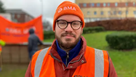 A man with brown facial hair and black glasses with rain on the lenses. He is wearing an orange beanie with white writing that says "BMA". He is wearing a red waterproof jacket and an orange and silver hi-vis jacket on top.