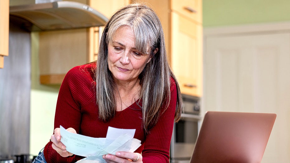 A mature woman leans on her kitchen counter in her Newcastle upon Tyne home, looking over bills and invoices beside an open laptop.