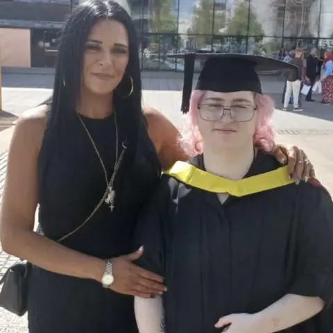 Lianne Hannam Two woman stood in front of a glass building. One has died pink hair and is wearing a black graduation cap and gown. The other has long dar hair and wears a back dress and cross body bag.