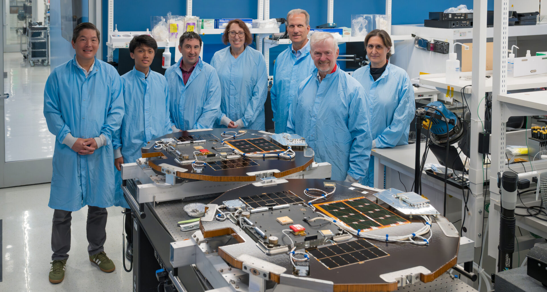 Seven people wearing blue lab coats stand near a lab table that supports two disk-shaped spacecraft