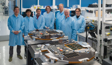 Seven people wearing blue lab coats stand near a lab table that supports two disk-shaped spacecraft