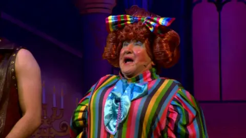 John singing on stage as May McFetridge in rainbow coloured outfit. 