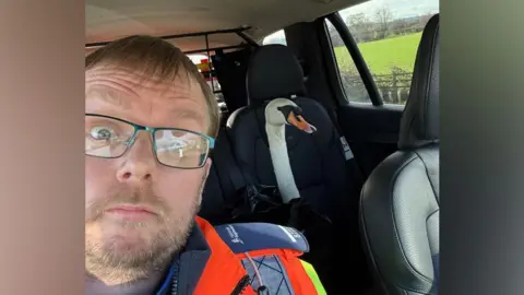 National Highways A selfie of Adrian Woods in high-vis traffic officer uniform poses in a car with a swan that has been strapped into a bag so it can be taken to safety.