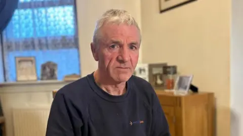 Alan Love, who has grey hair, is sitting at a table. He is wearing a dark, long-sleeve top. The background includes a window with patterned curtains, framed photos, a lamp, and a wooden cabinet. 