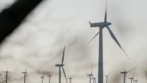 Images showing the top sections of several wind turbines spinning against a grey sky