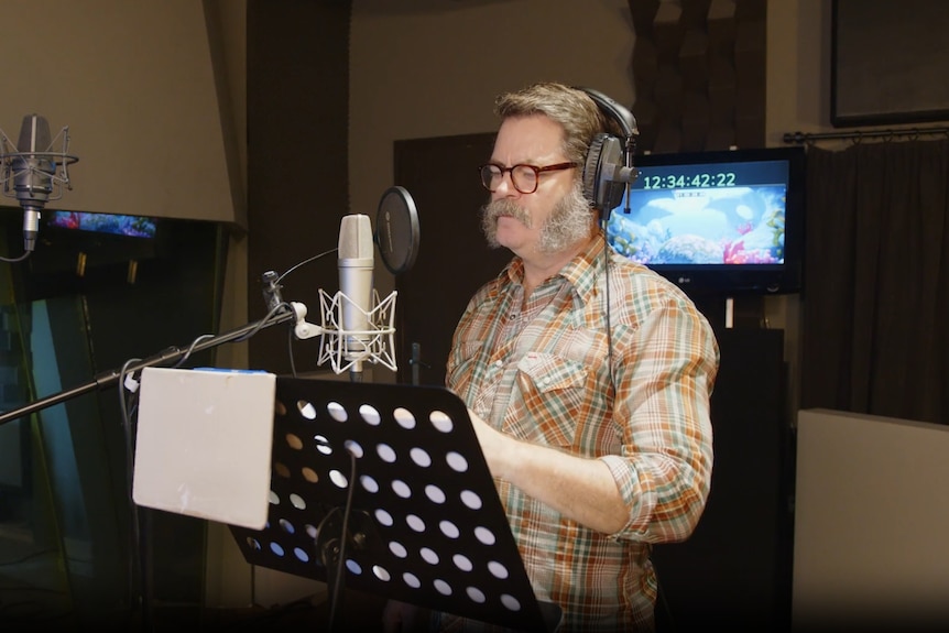 Nick Offerman with reading glasses and moustache wearing headphones and speaking into a mic