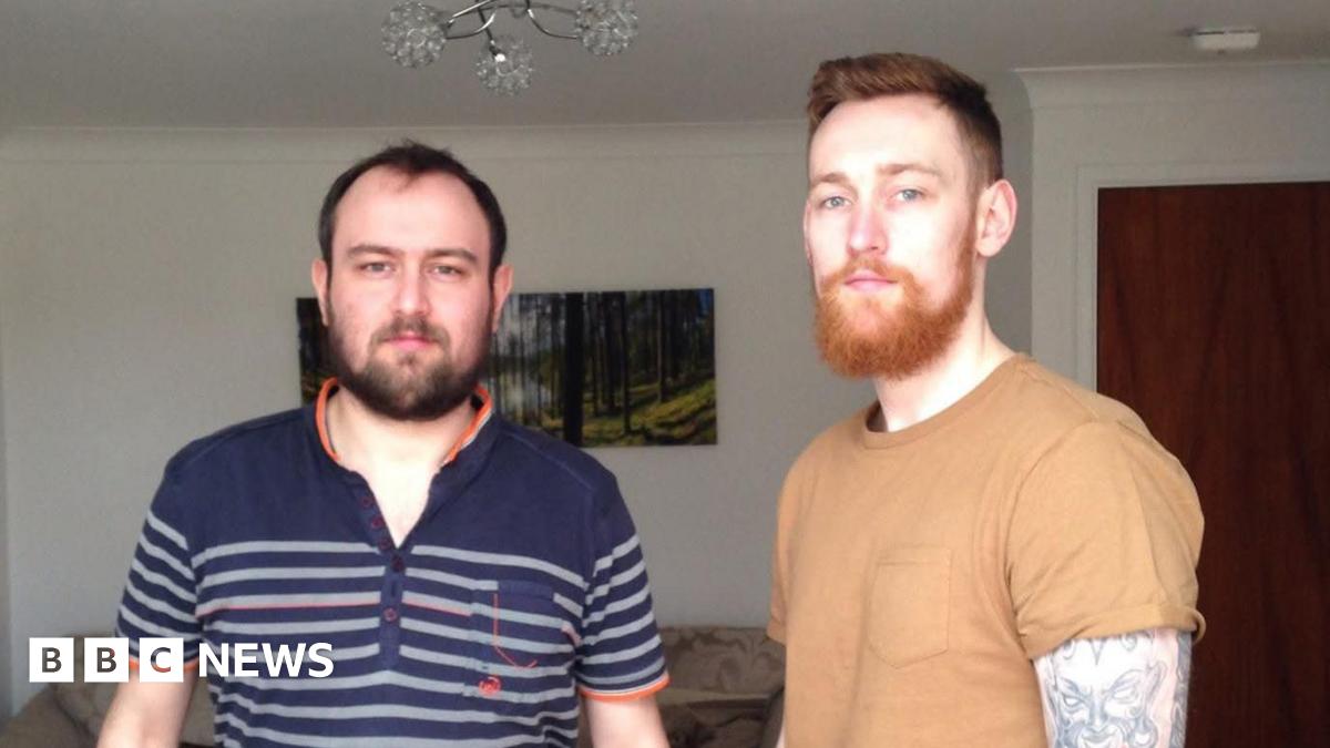Two men stood in a living room. The man on the left has black hair, black beard, navy polo t-shirt with orange trim and grey stripes. The man on the right has red hair, a red beard, a light brown t-shirt and a heavily tattooed left arm. The living room is cream, with a cream settee, woodland photograph, dark wooden door and a flowery light fitting.